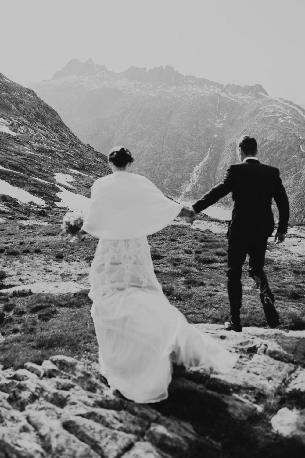 Black and white photo of a couple holding hands while walking on a mountain ridge.