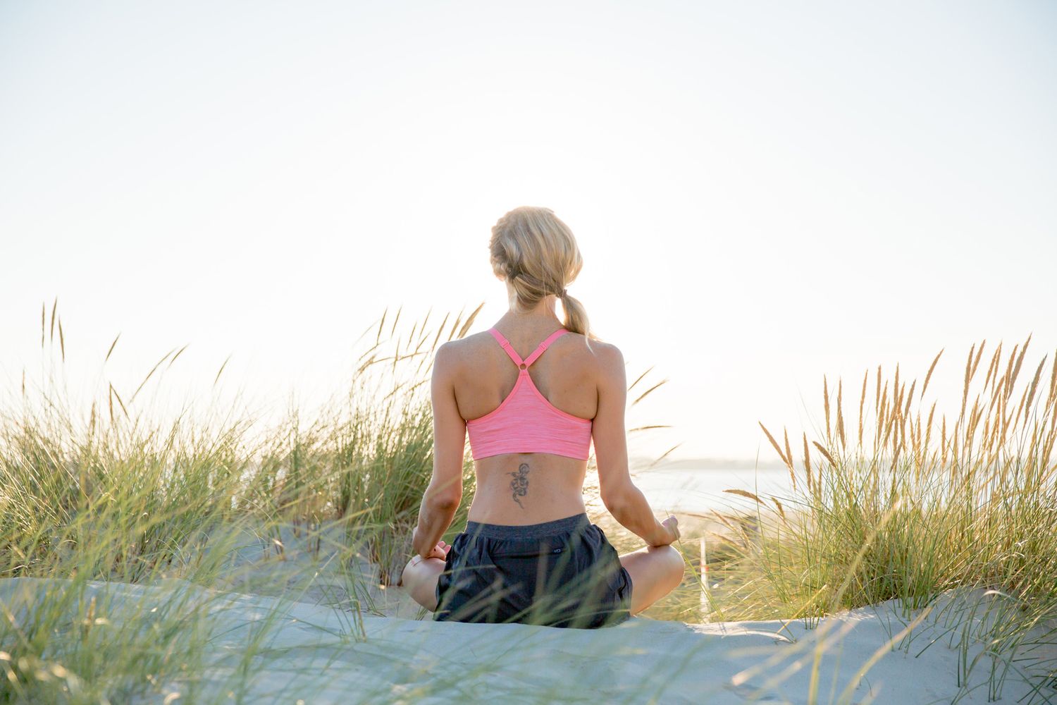 Person in pink sports top sitting peacefully among beach grass at sunset during business branding photoshoot.