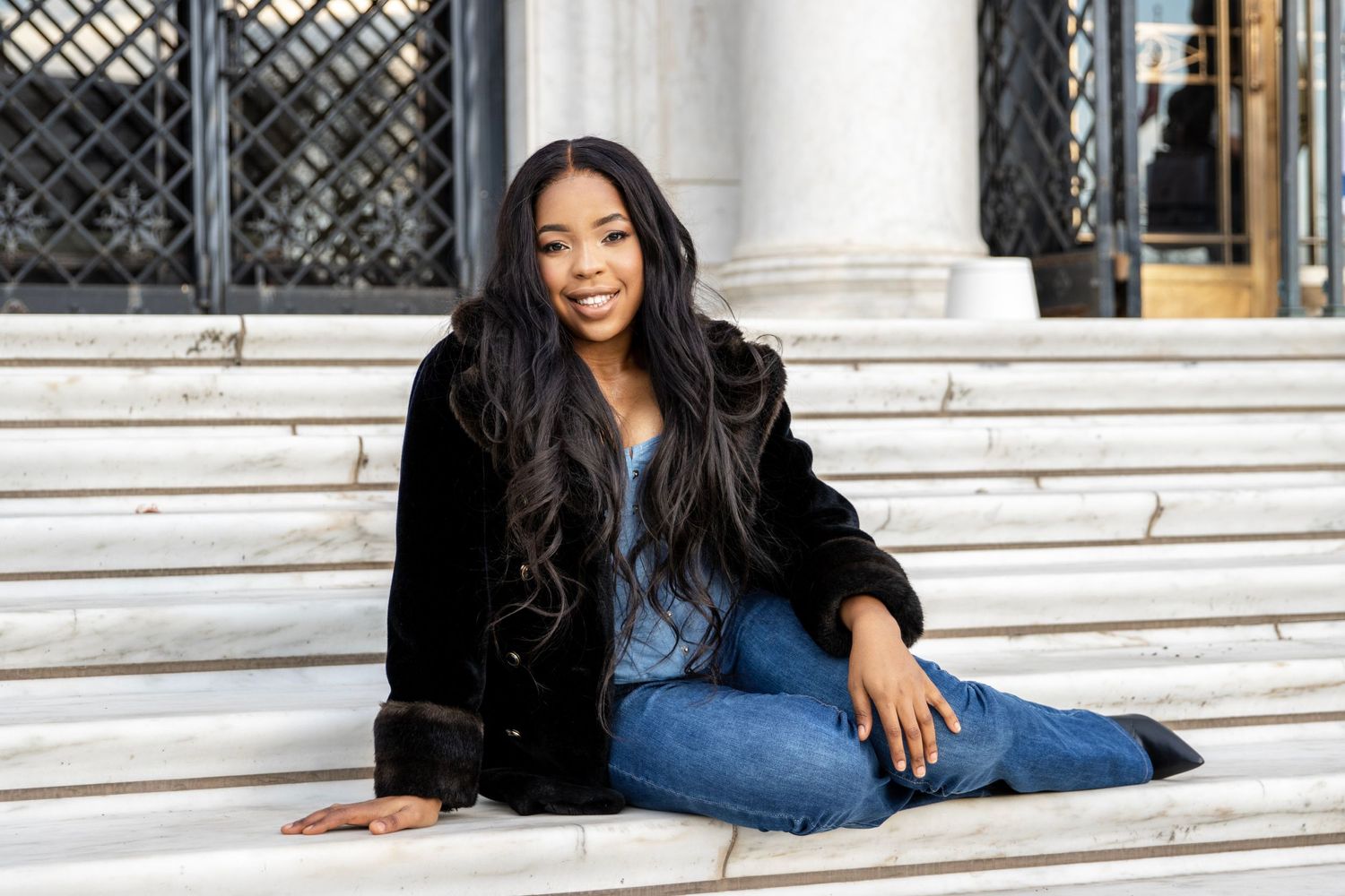 A woman in a black fur coat and blue jeans sits casually on white marble steps outside of the detroit institute of arts