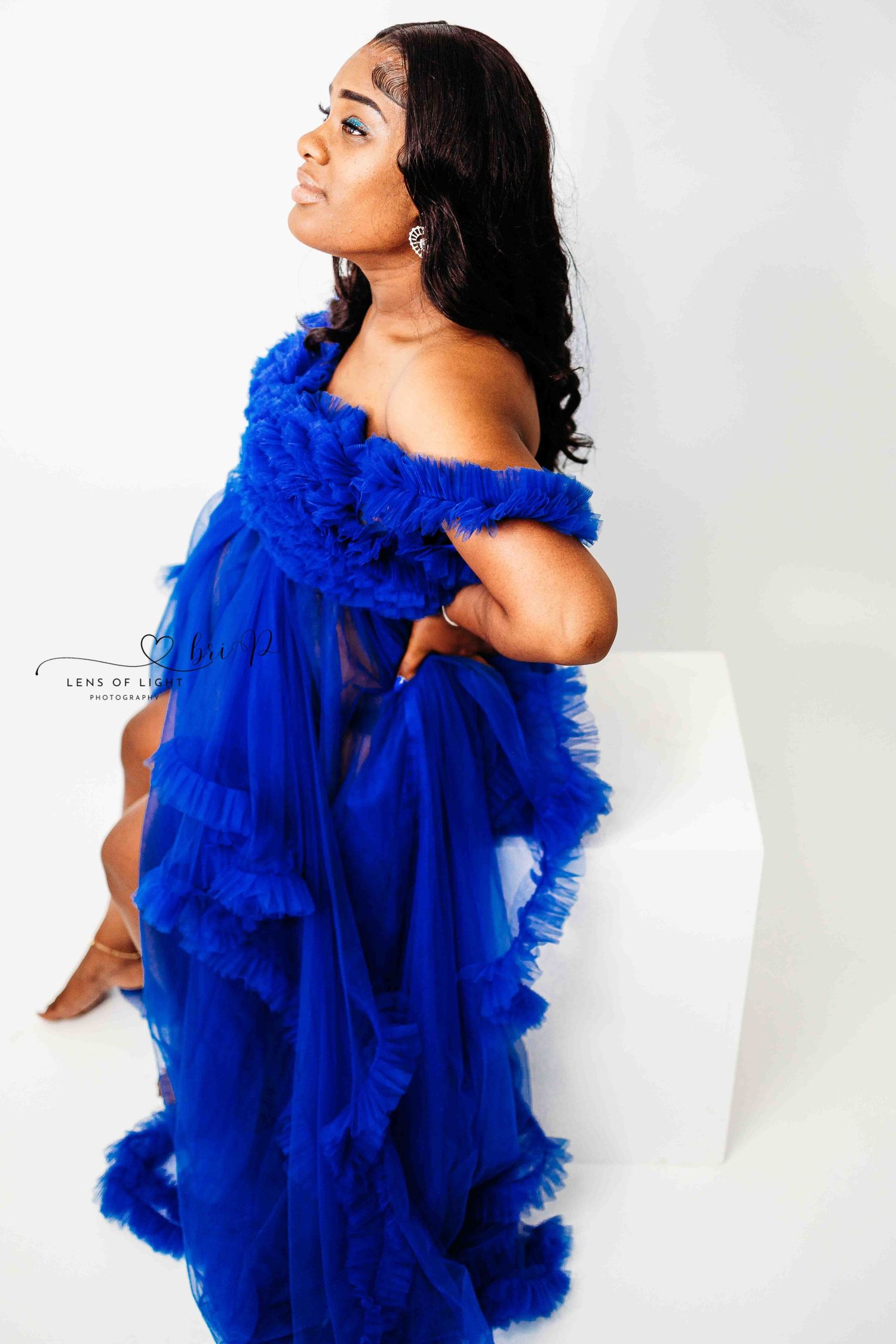 Side profile of woman in flowing royal blue tulle gown against white studio backdrop.