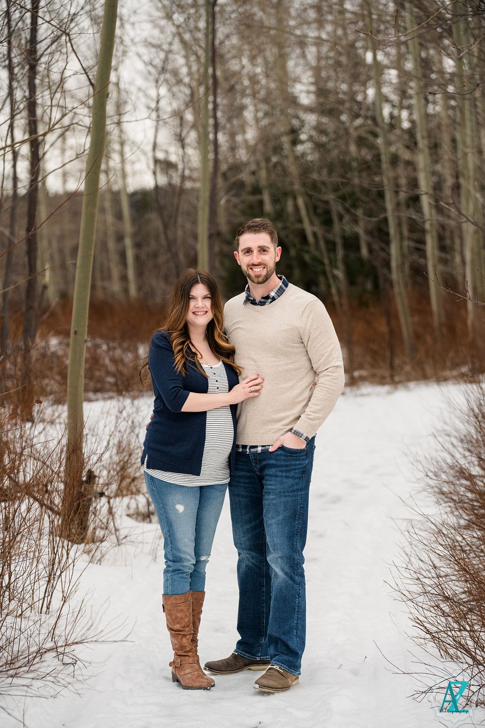 Winter engagement photo of couple in casual wear standing in snowy wooded area.
