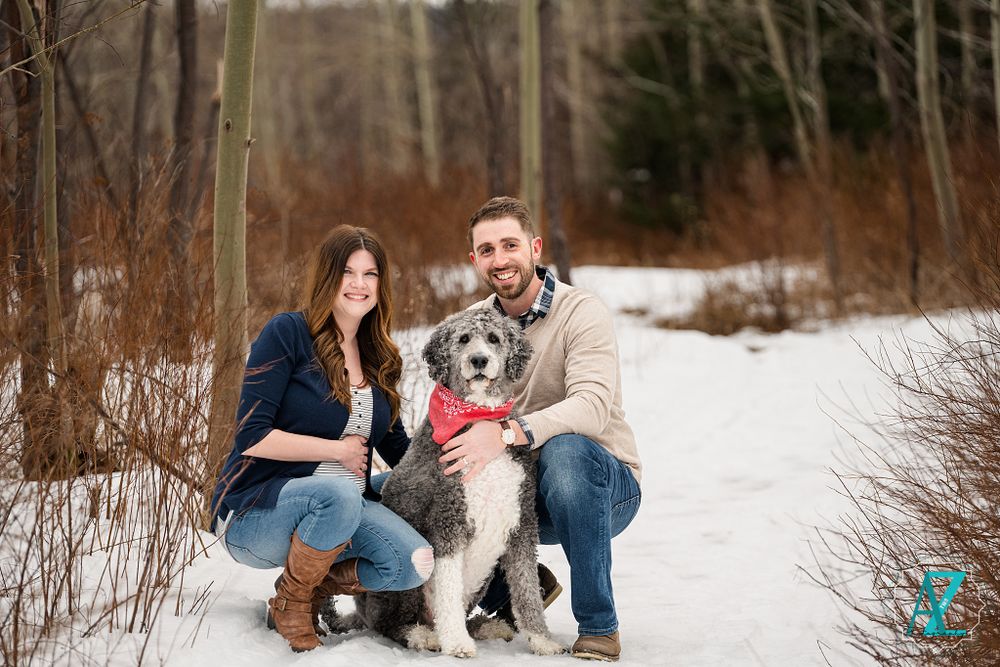 Casual winter portrait of couple with their dog in snow-covered forest setting.