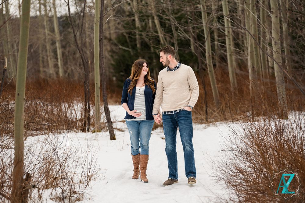 A couple walks together on a snowy winter trail through bare trees wearing coordinating casual outfits.