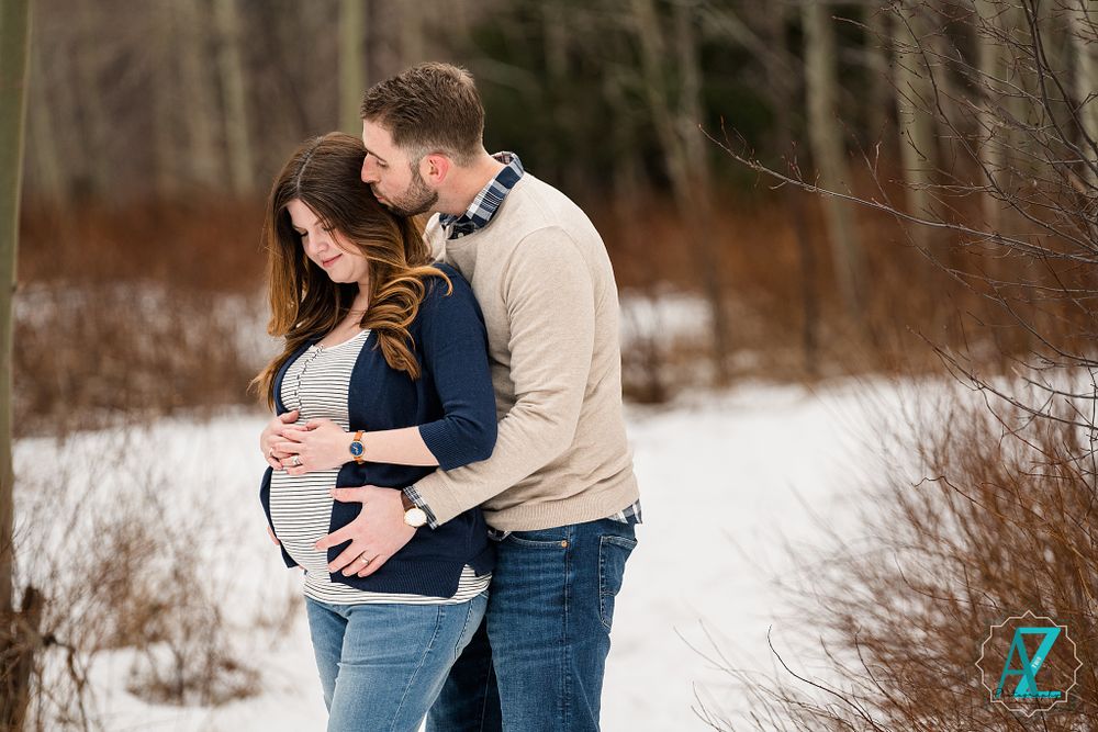 Intimate maternity portrait of couple embracing in snowy winter forest setting.