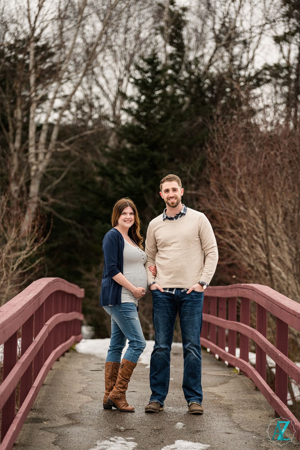 Expectant couple poses together on red wooden bridge during winter photo session.
