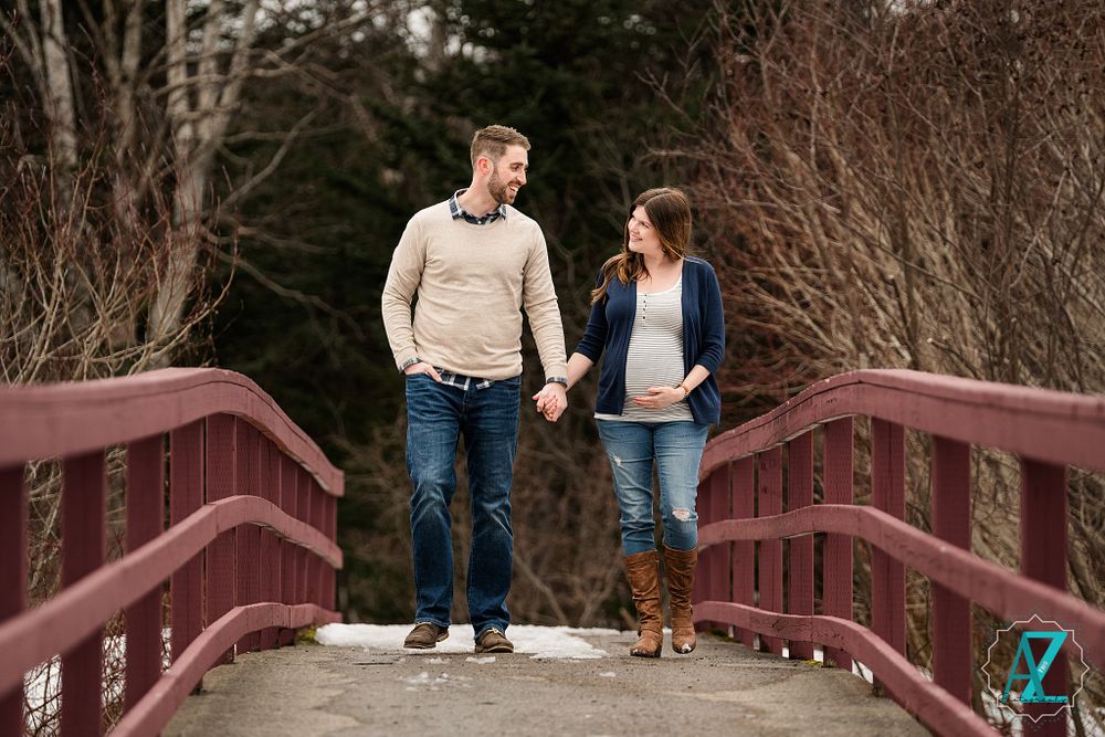 Couple holds hands while standing on rustic red bridge during winter maternity photoshoot.