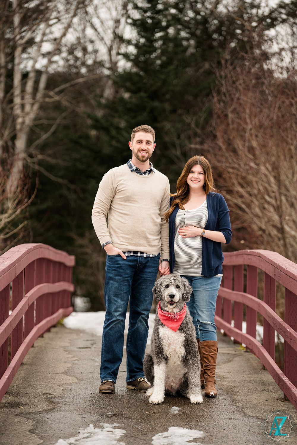 Couple and their shaggy dog with red bandana pose together on wooden bridge in winter setting.