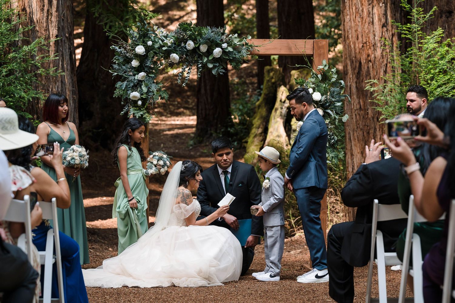 An outdoor wedding ceremony in a redwood grove with a floral arch and seated guests.