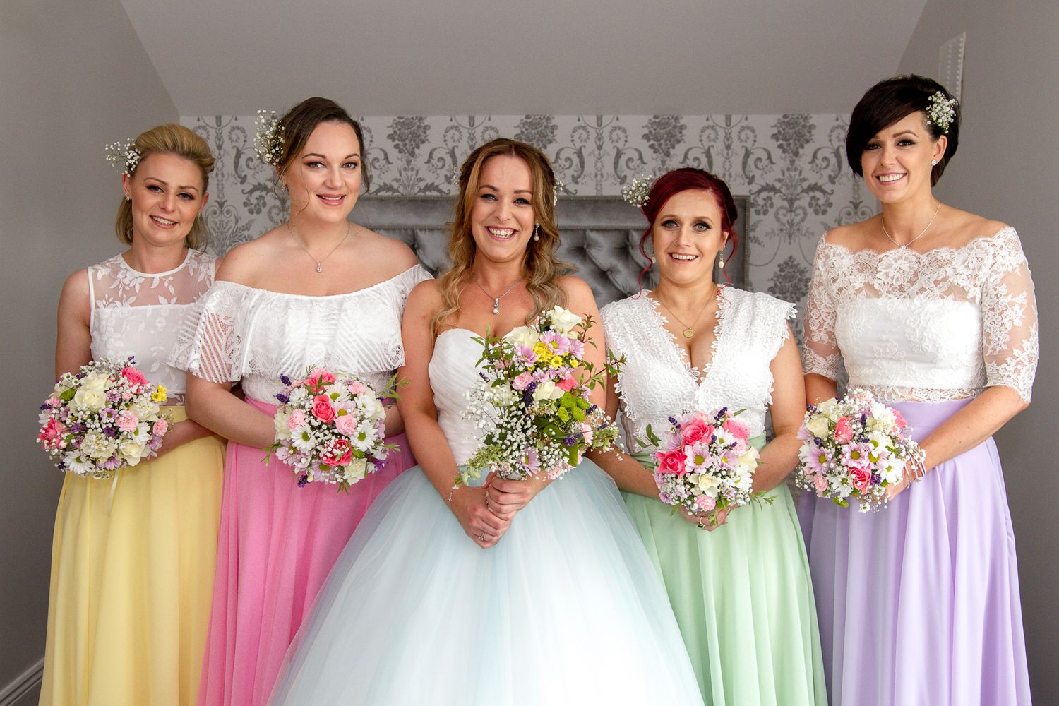 Bridesmaids in pastel colored dresses holding floral bouquets pose together at a wedding.