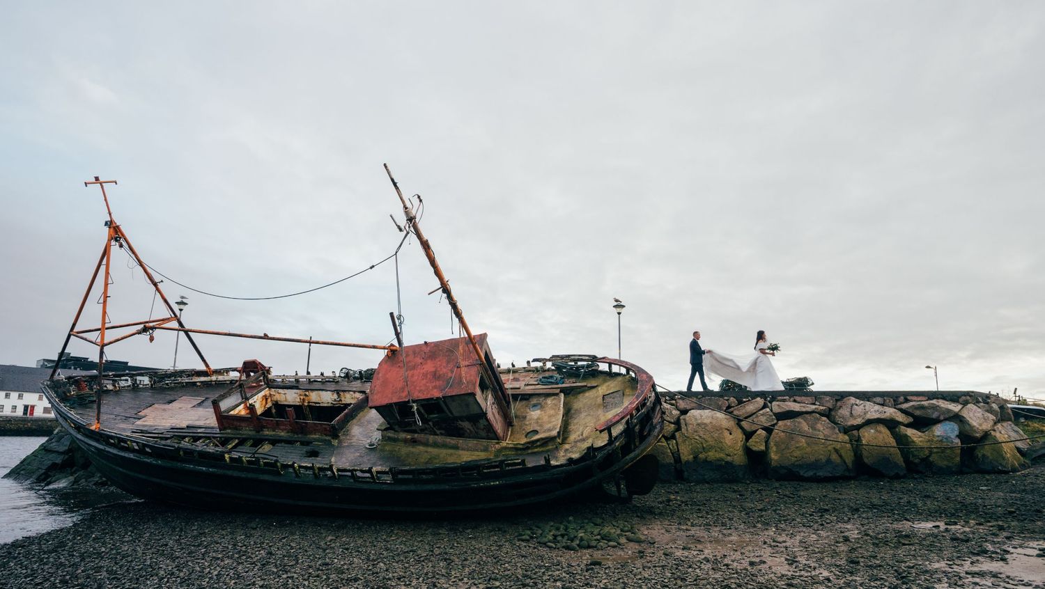 An old weathered fishing boat rests against a rocky breakwater under an overcast sky.