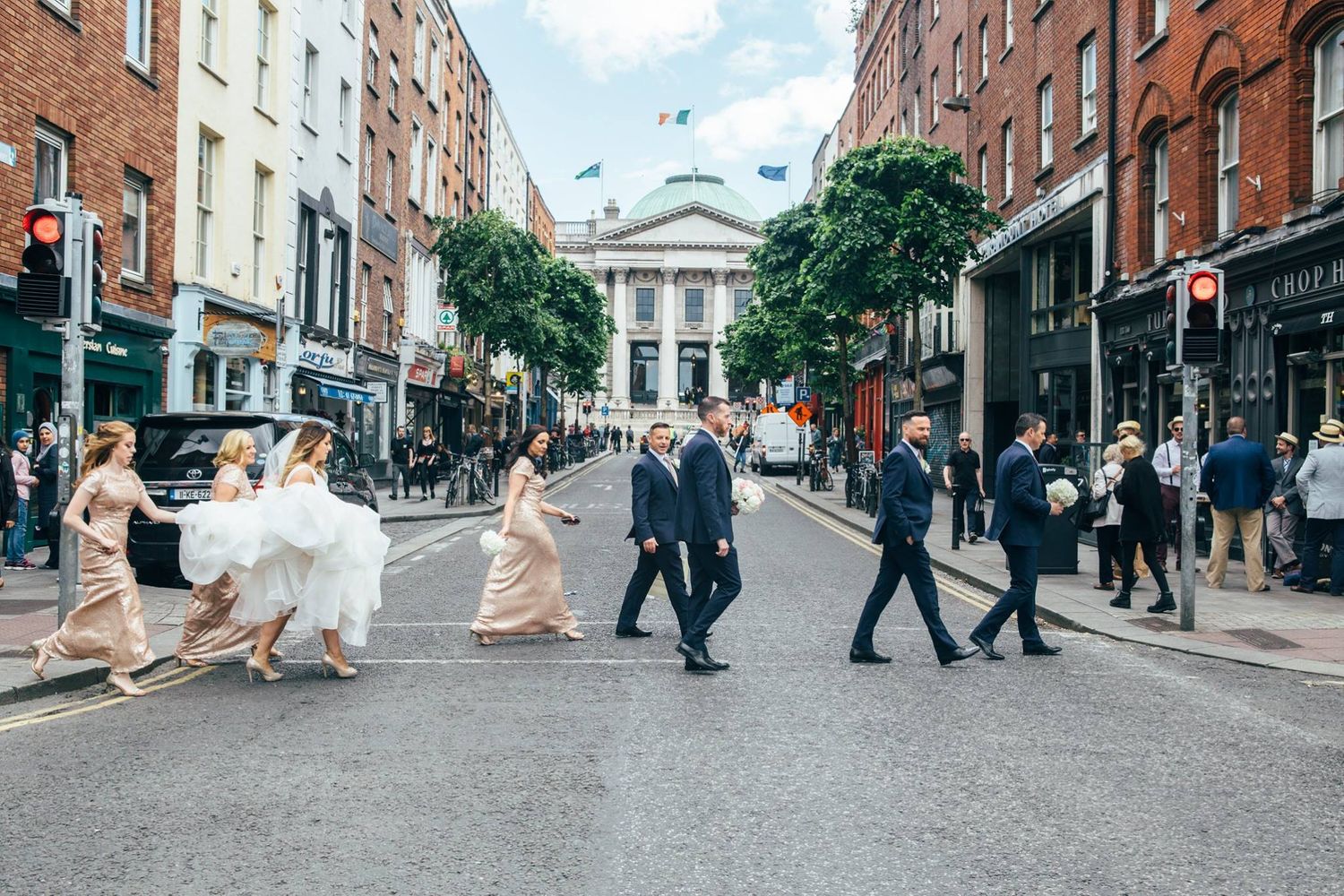 Wedding party walking across urban street with historic architecture in background.