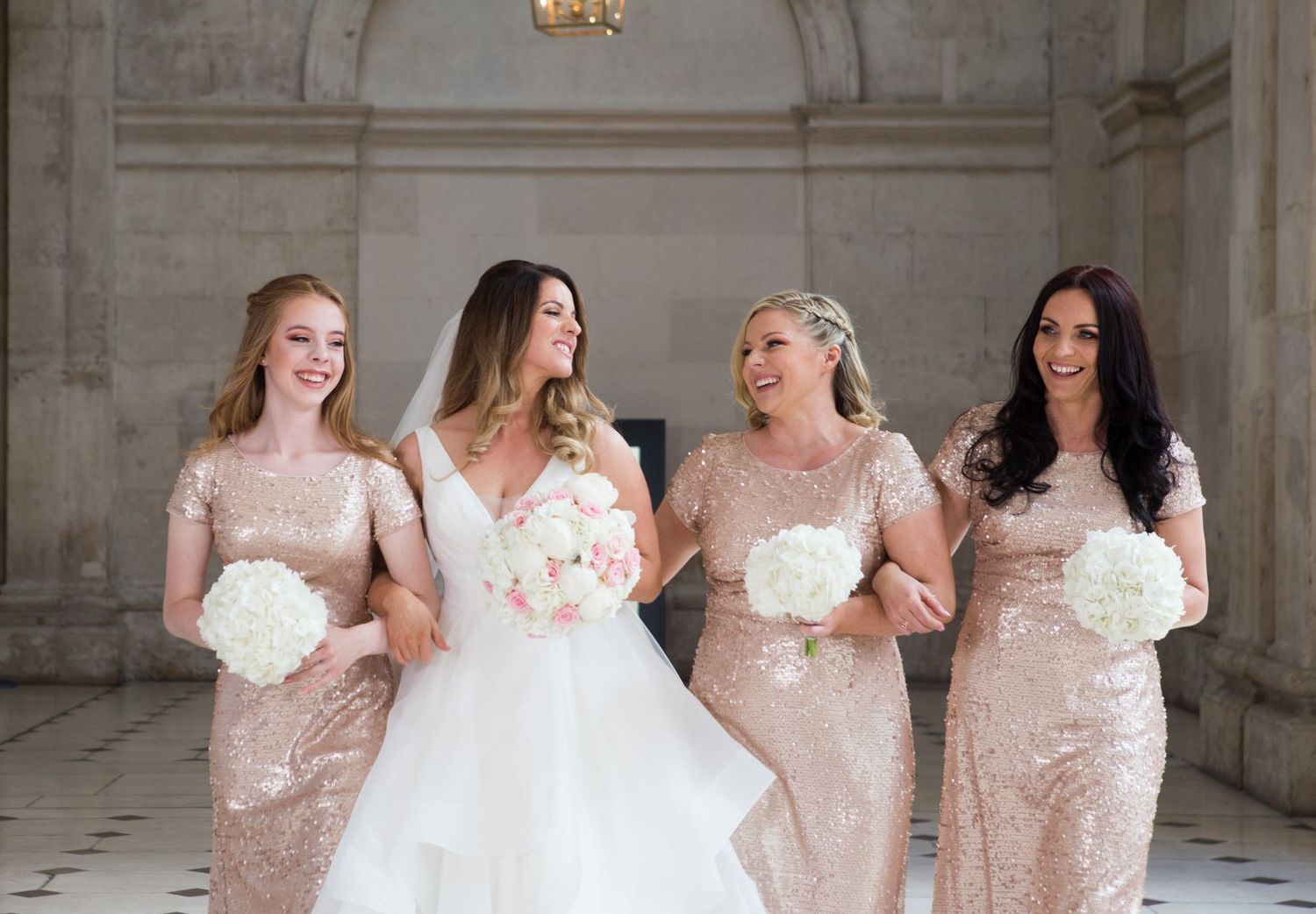 Bridesmaids in blush pink dresses pose together in an elegant indoor venue with white columns.