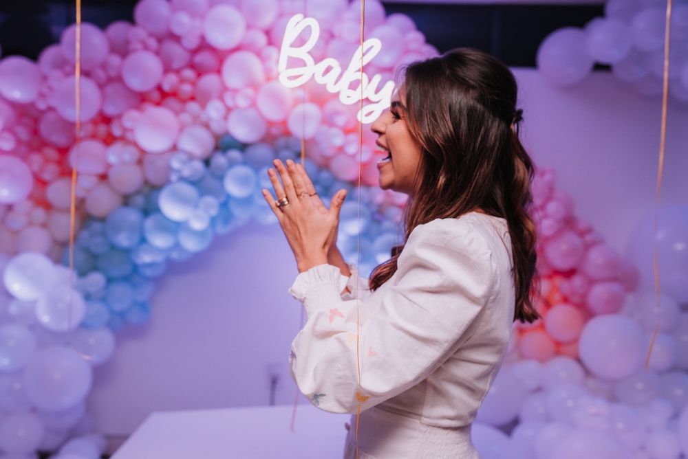 Woman in white outfit expressing excitement in front of pink and purple balloon arch with Baby sign.