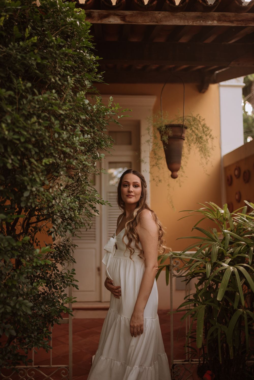 Expectant mother in white dress poses under rustic porch with lush greenery.