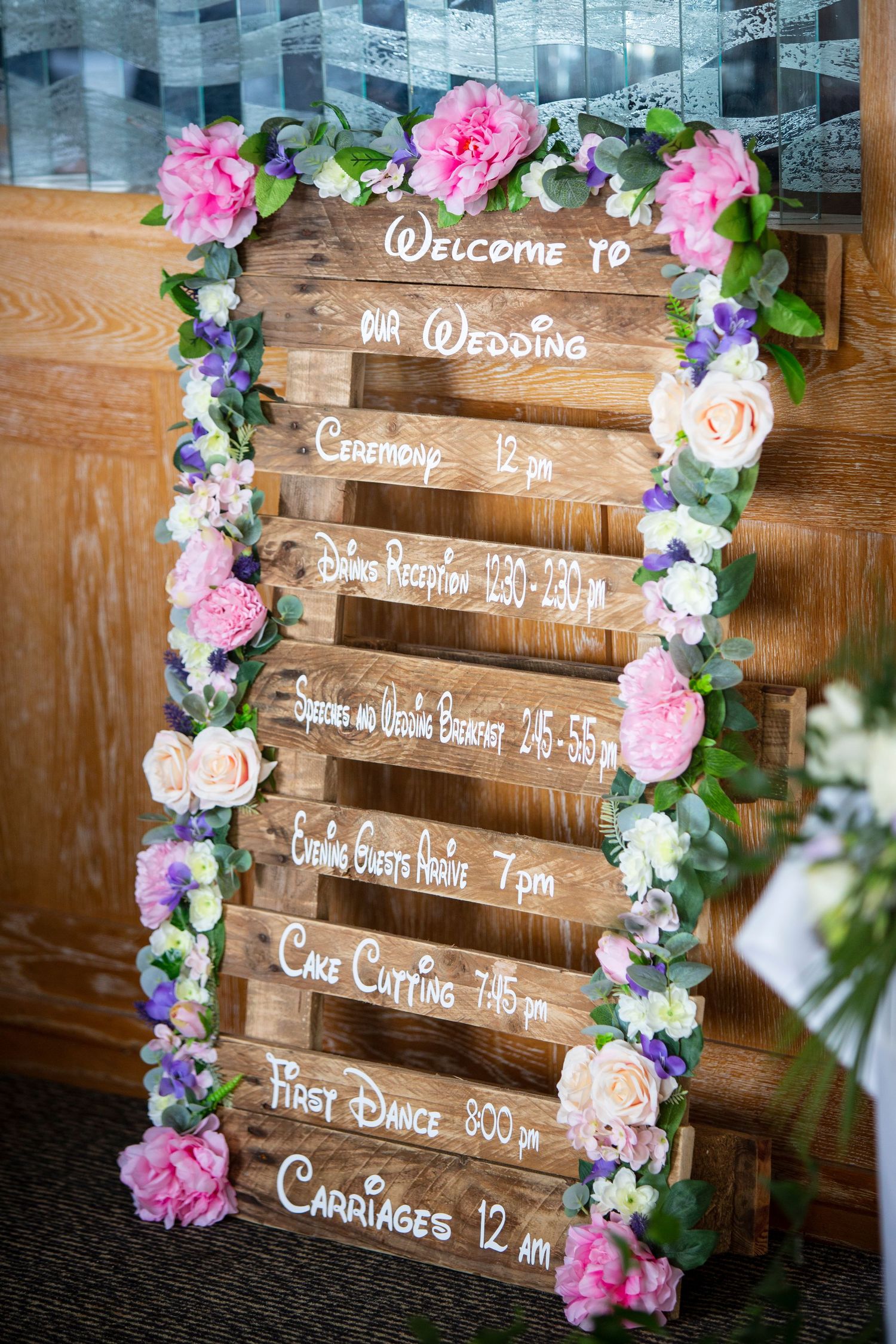Rustic wooden wedding timeline sign decorated with pink and purple floral garland.