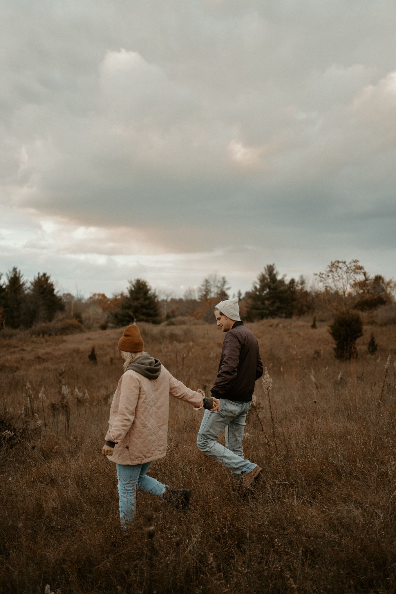 Couple holding hands walking through an autumn field under cloudy skies, wearing winter hats and casual outdoor clothing.
