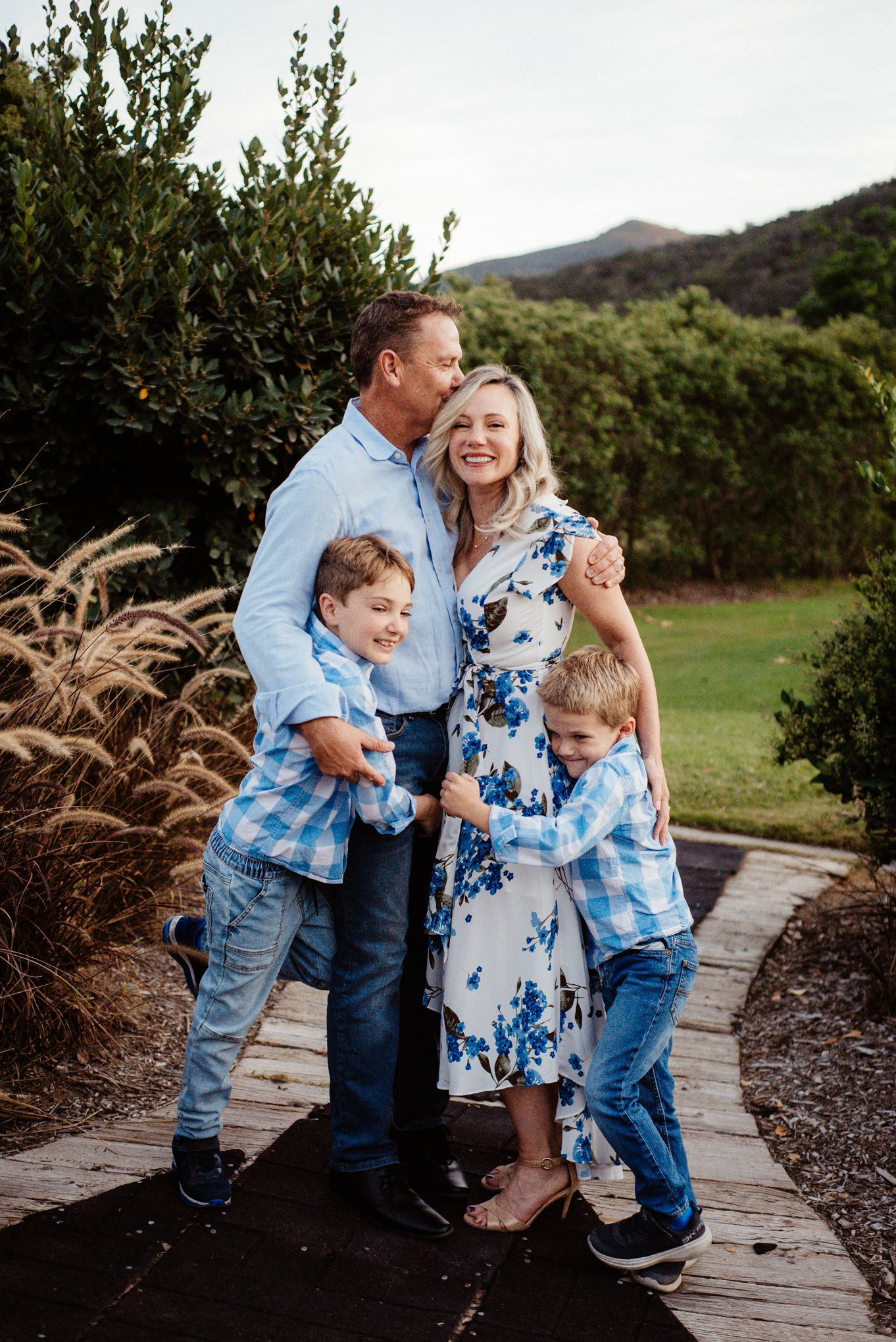 A family in coordinating blue and white outfits poses together on a wooden pathway outdoors.