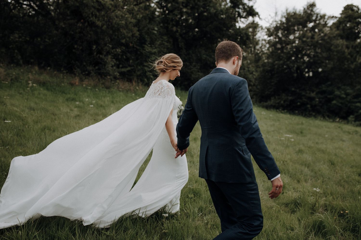 Bride in a flowing cape gown walks hand in hand with groom across a green hillside.