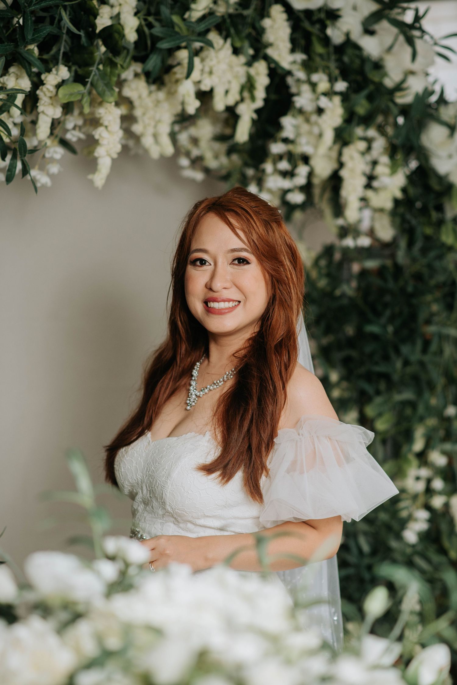 Bride in off-shoulder wedding dress poses against backdrop of cascading white flowers.