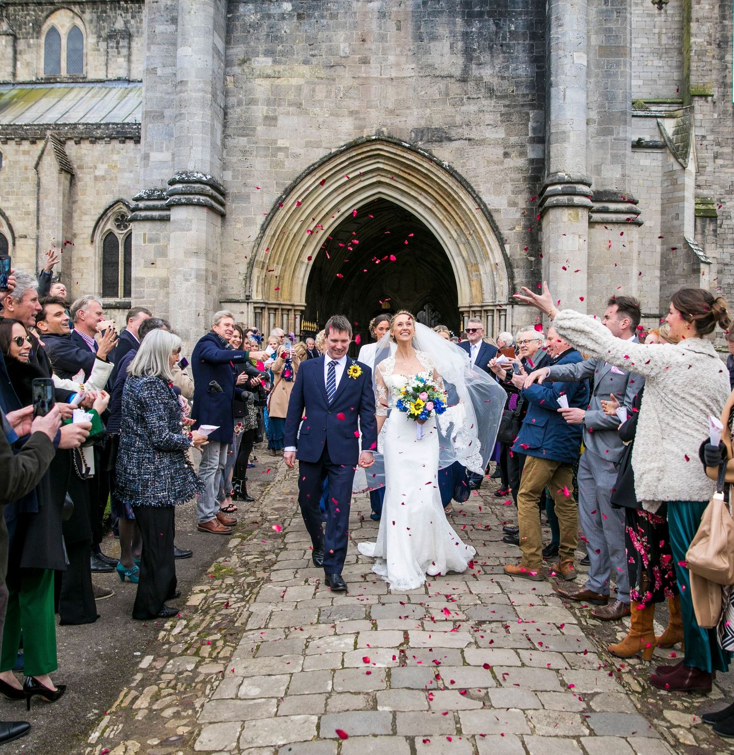 Wedding guests line a church pathway throwing petals as newlyweds exit the ceremony.