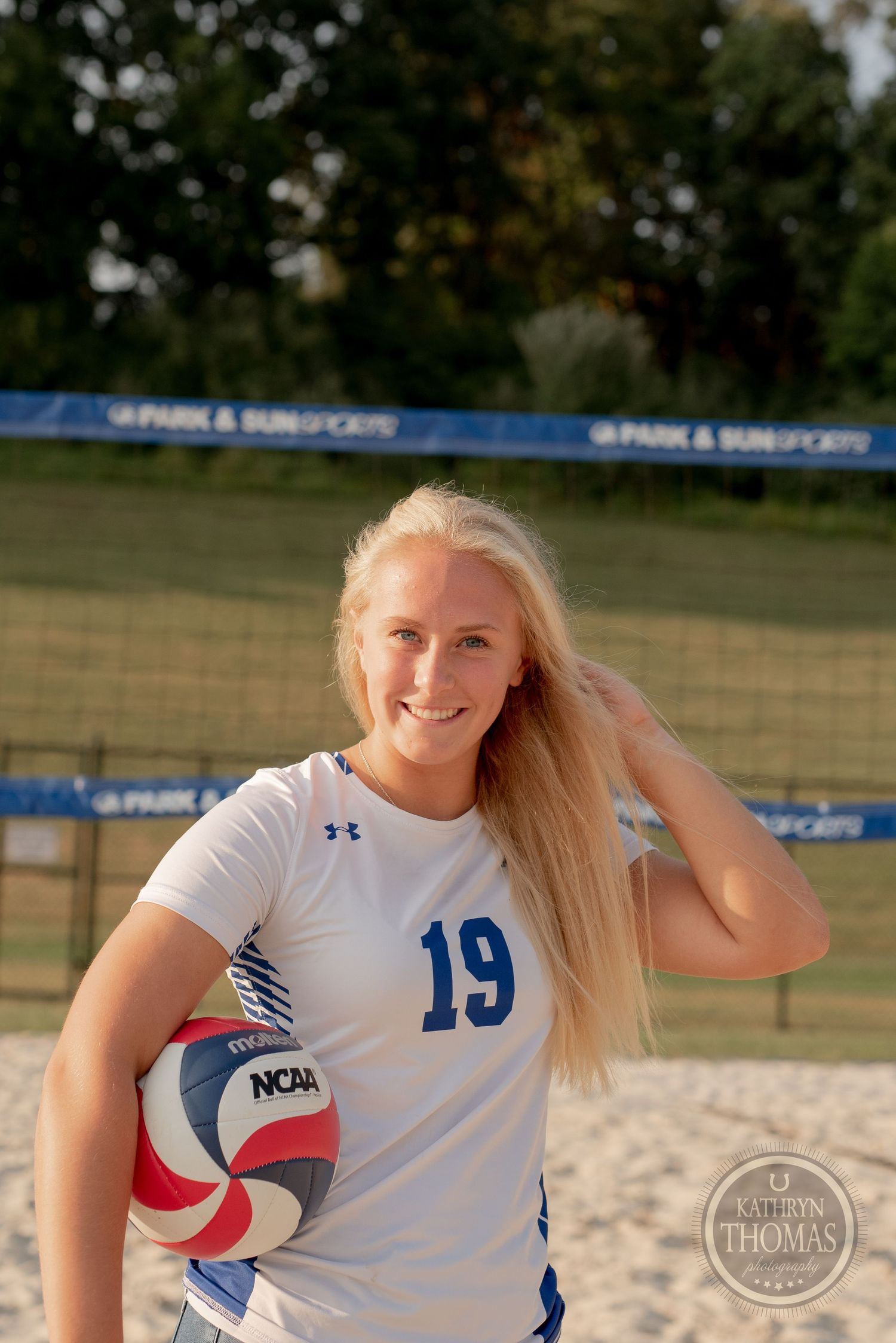 An athlete in jersey 19 poses with a volleyball on a sunny beach court.