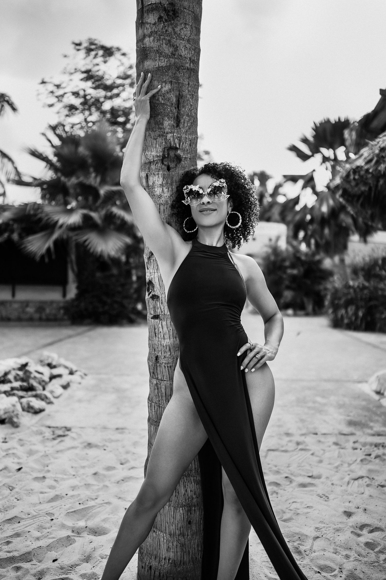 Stylish black and white photo of person in high-slit dress posing against palm tree on beach.