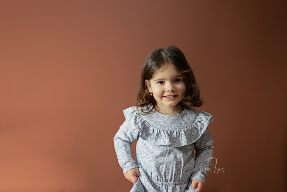 Smiling toddler girl in a light blue ruffled dress posing against a warm brown studio background.