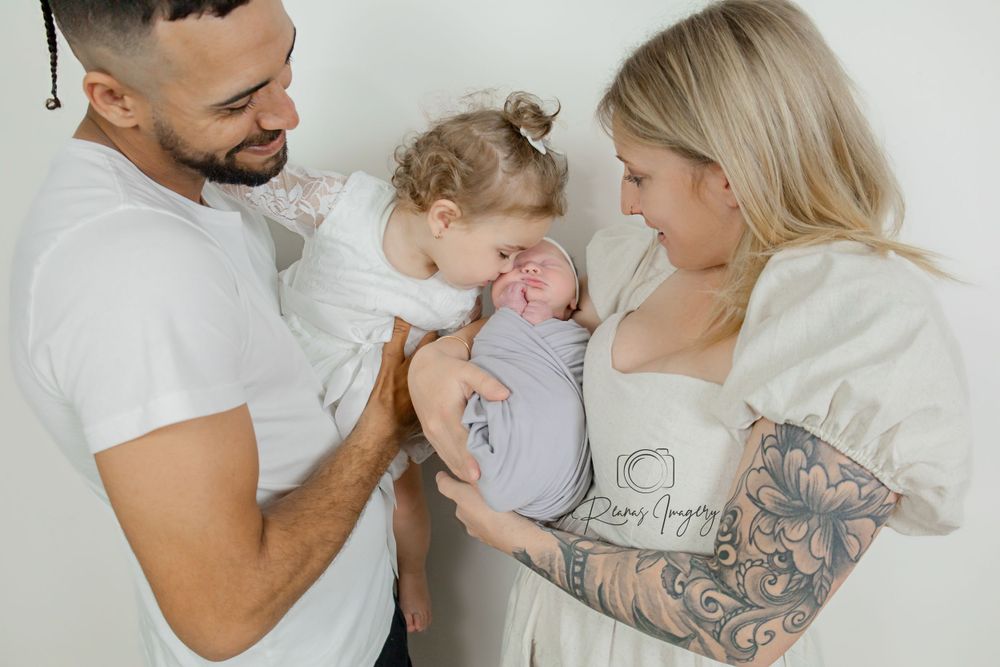 Happy family of three with tattooed mother and father as toddler kisses their newborn baby.