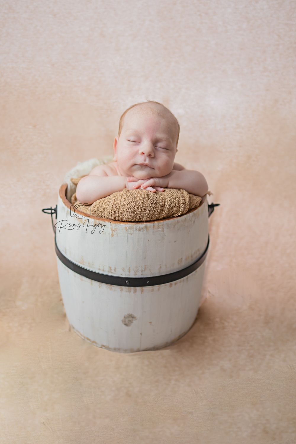 Peaceful sleeping newborn baby posed in a rustic white wooden bucket with a golden knit cushion.