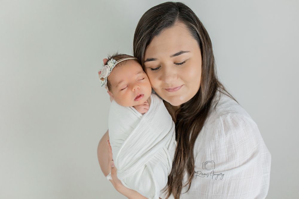 A mother tenderly holds her swaddled newborn baby against a soft white studio background.