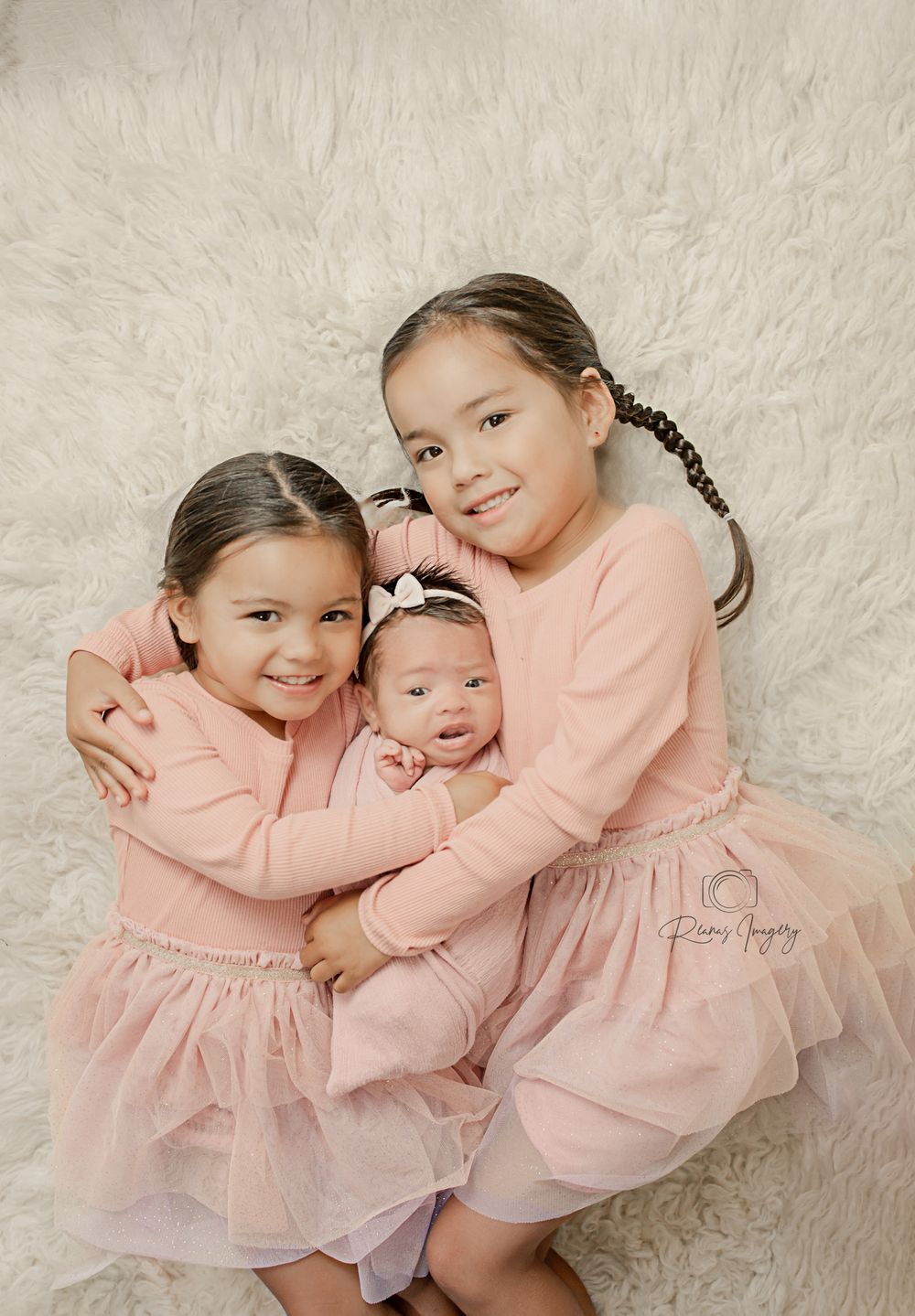 Two young sisters in matching pink outfits lovingly hold their newborn baby sibling on a white rug.