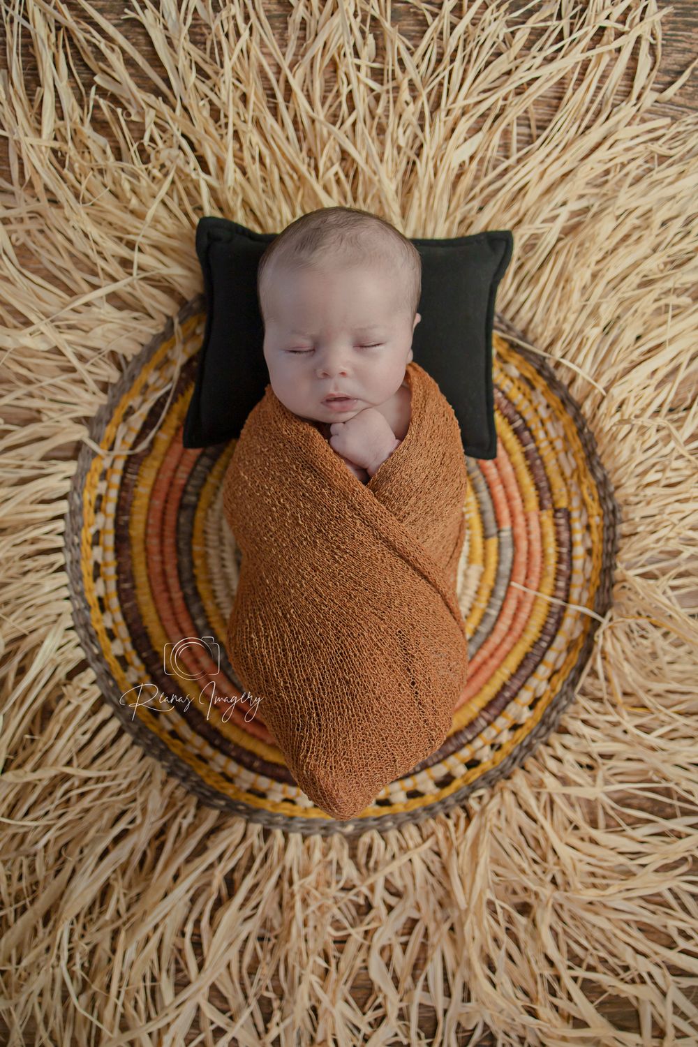 Sleeping newborn baby swaddled in orange wrap resting in a colorful woven basket on straw.