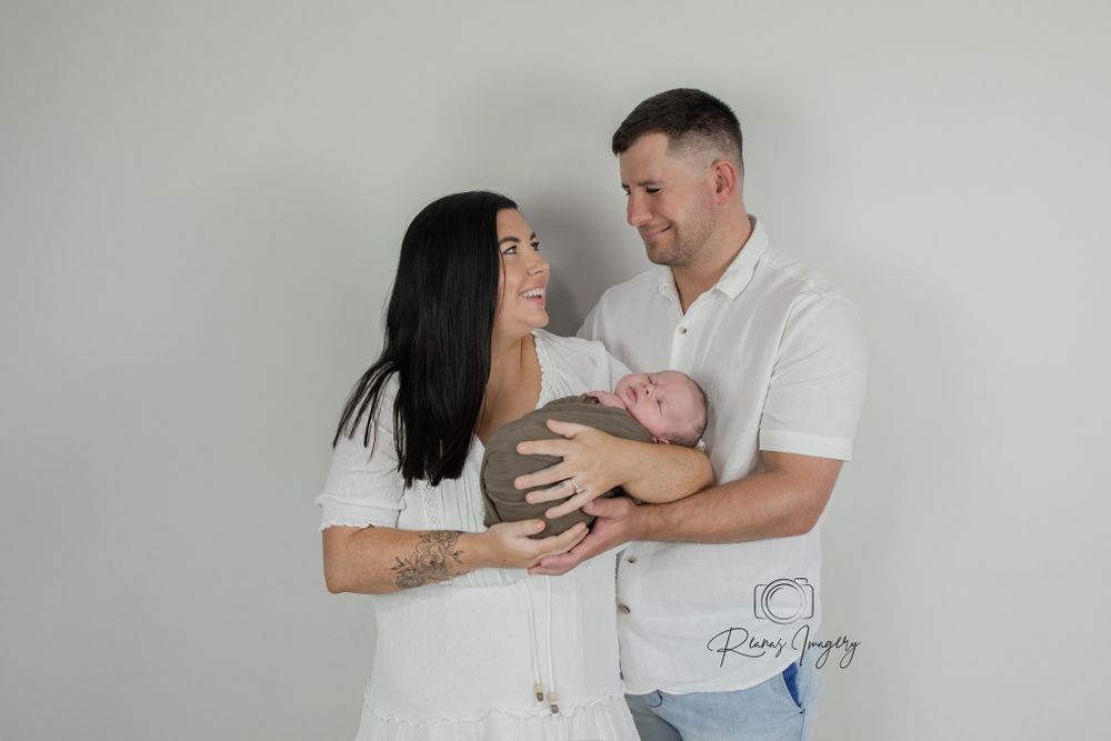 Smiling couple in white outfits proudly holds their newborn baby in a bright studio setting.