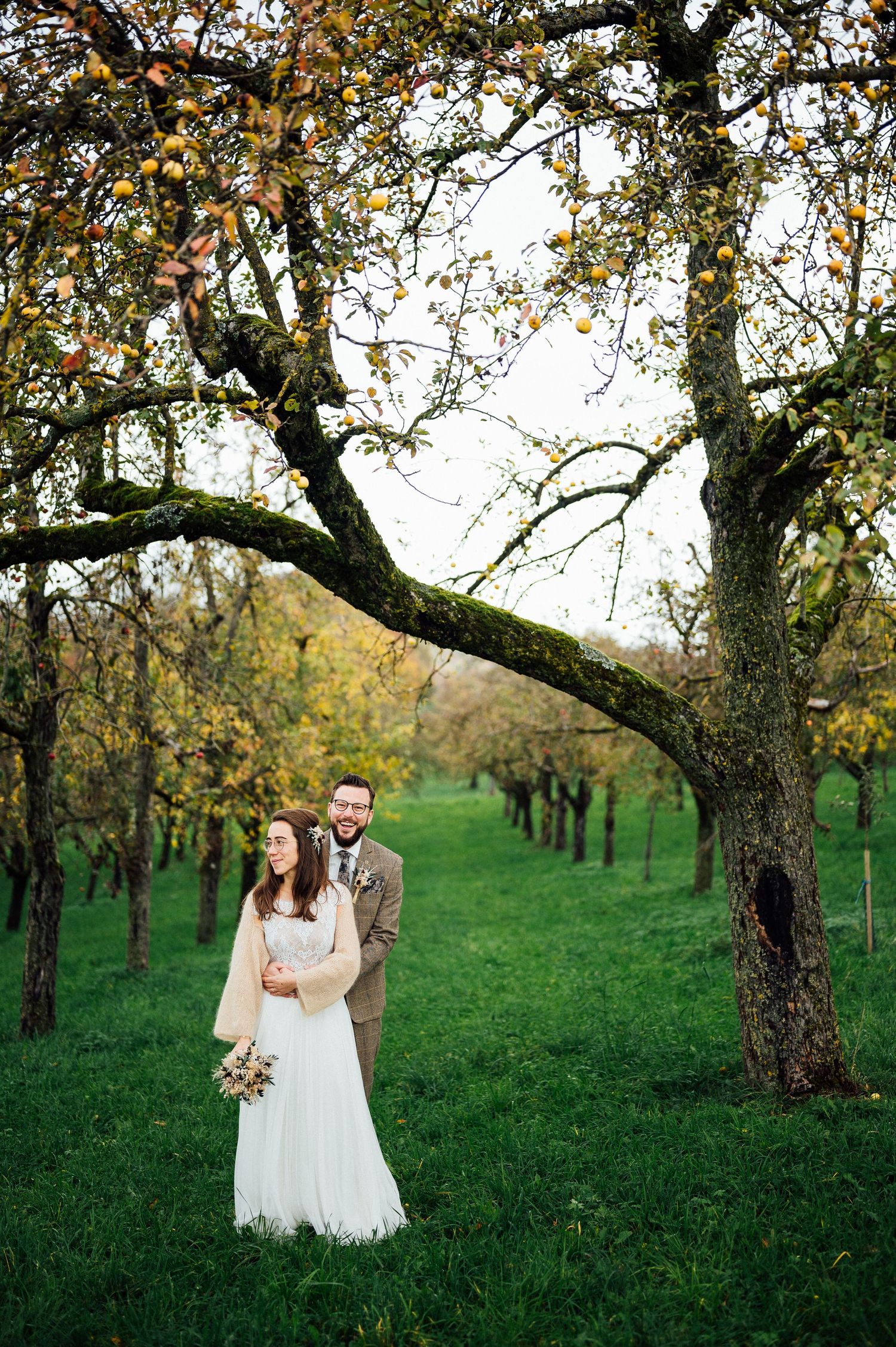 Couple standing in an orchard with autumn trees and green grass on their wedding day.