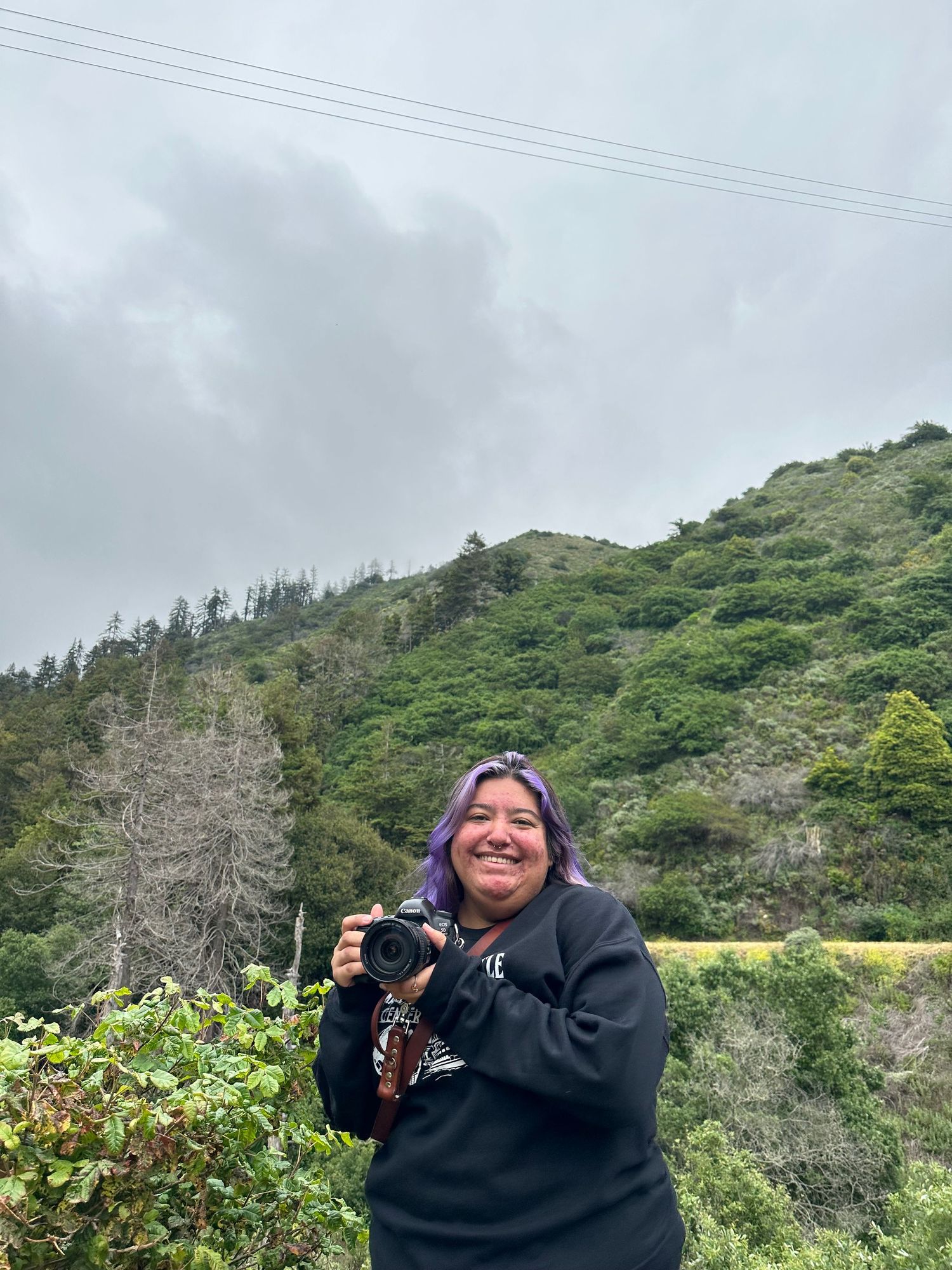 Person smiling while holding camera against green hillside and cloudy sky.