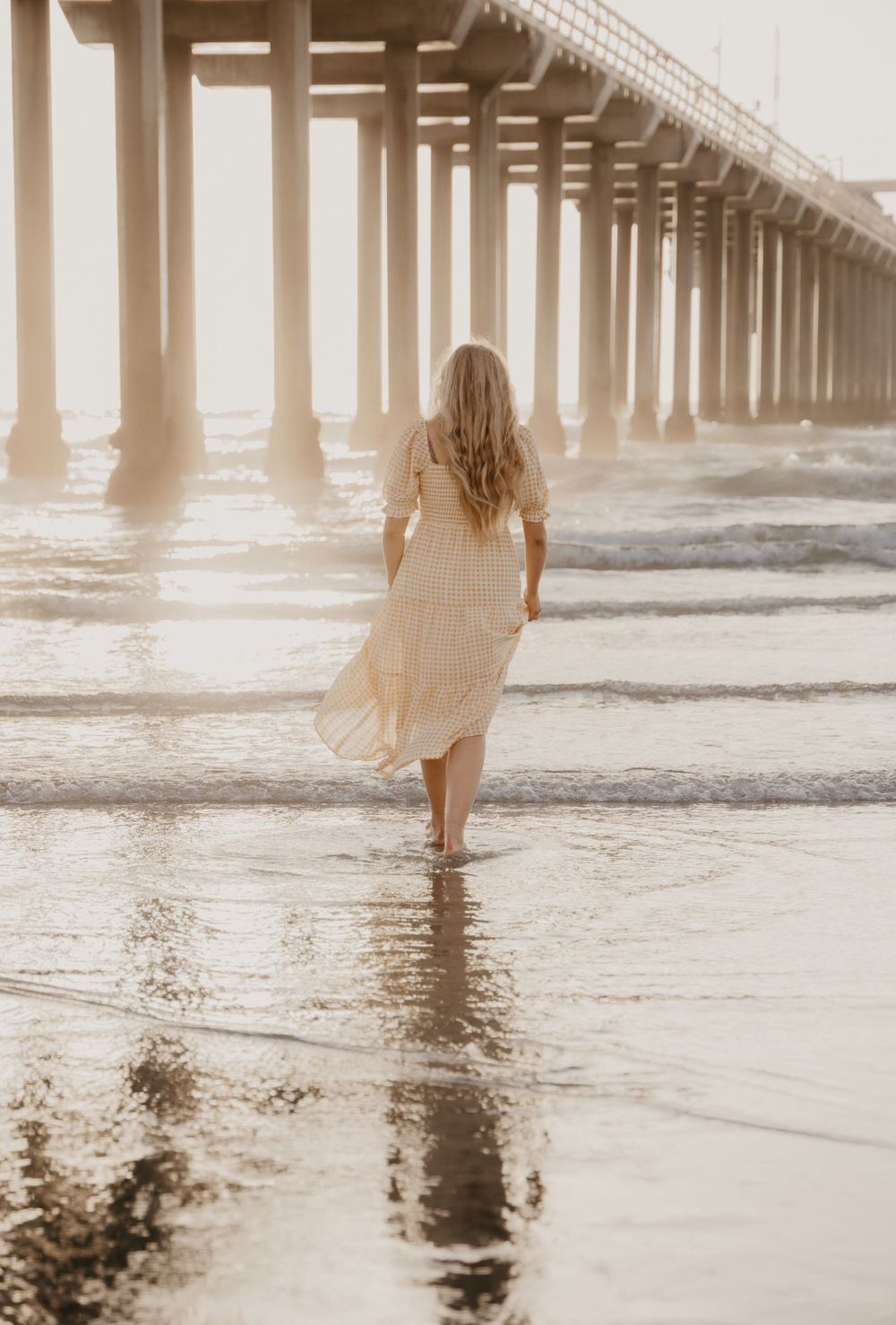Back view of person in flowing dress walking under pier at sunset.