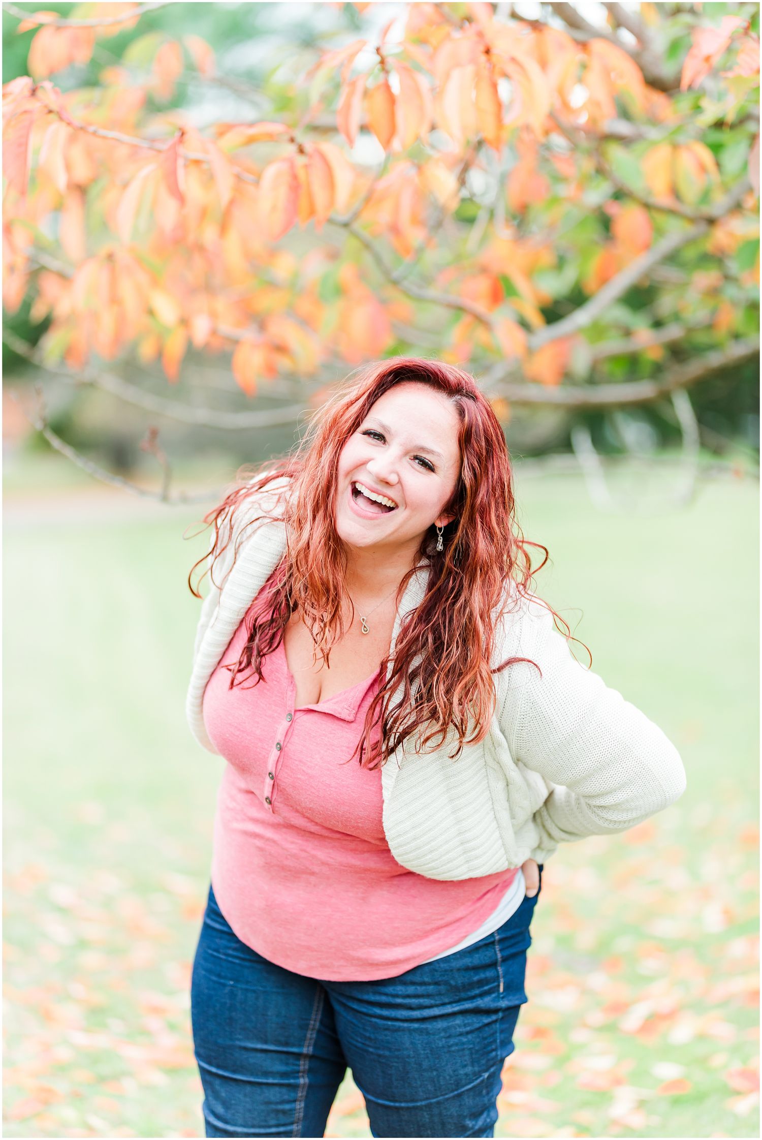 Person in pink top and white cardigan poses outdoors under orange autumn leaves.