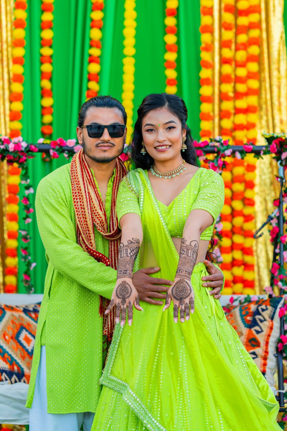 Couple in matching lime green traditional Indian outfits posing against marigold and floral decorations.