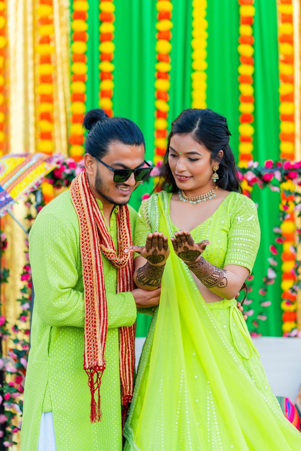 Couple in matching lime green attire showing henna designs against vibrant marigold backdrop.