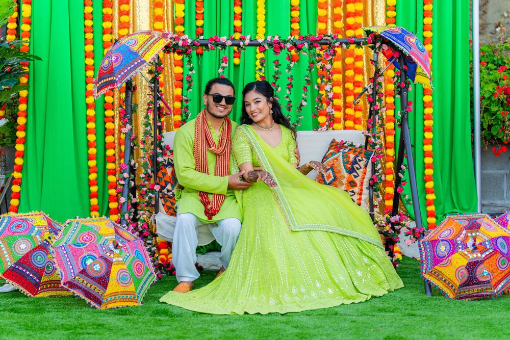 Traditional Indian couple in bright green attire posed with decorative umbrellas against marigold backdrop.