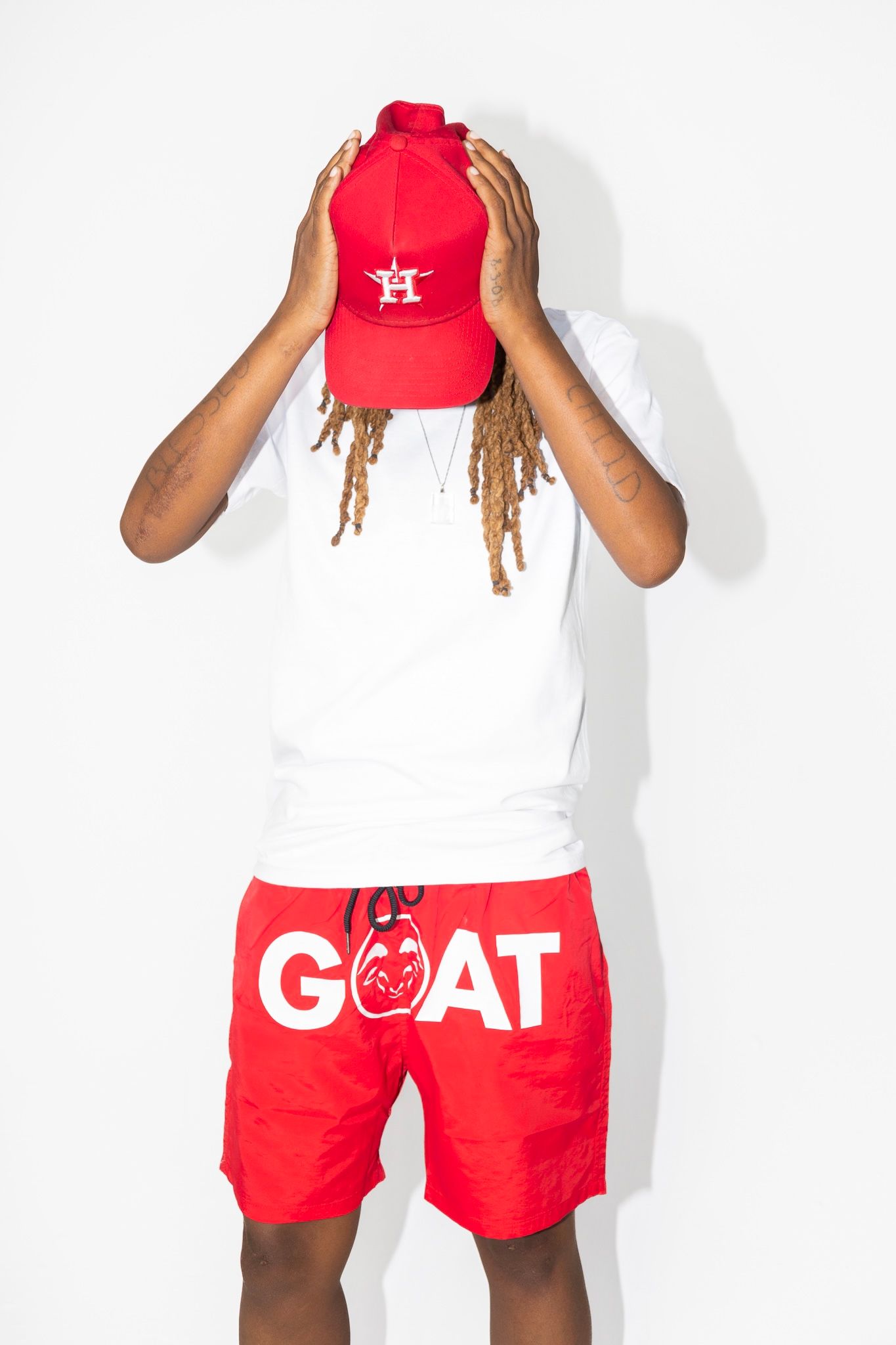 Person in white shirt and red GOAT shorts covering face with red baseball cap.
