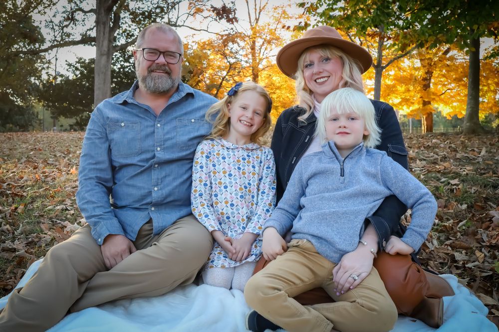 Family relaxing together on grass during golden hour with fall foliage in background.