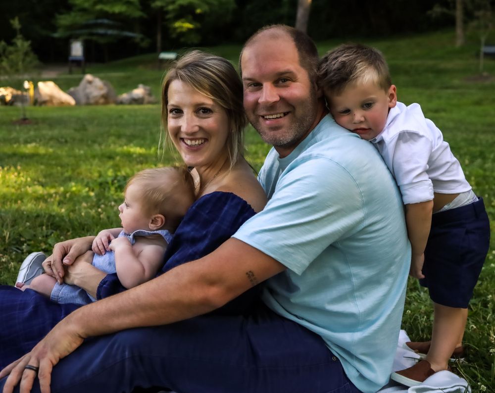 A happy family with two small children sits together on grass in a park setting.