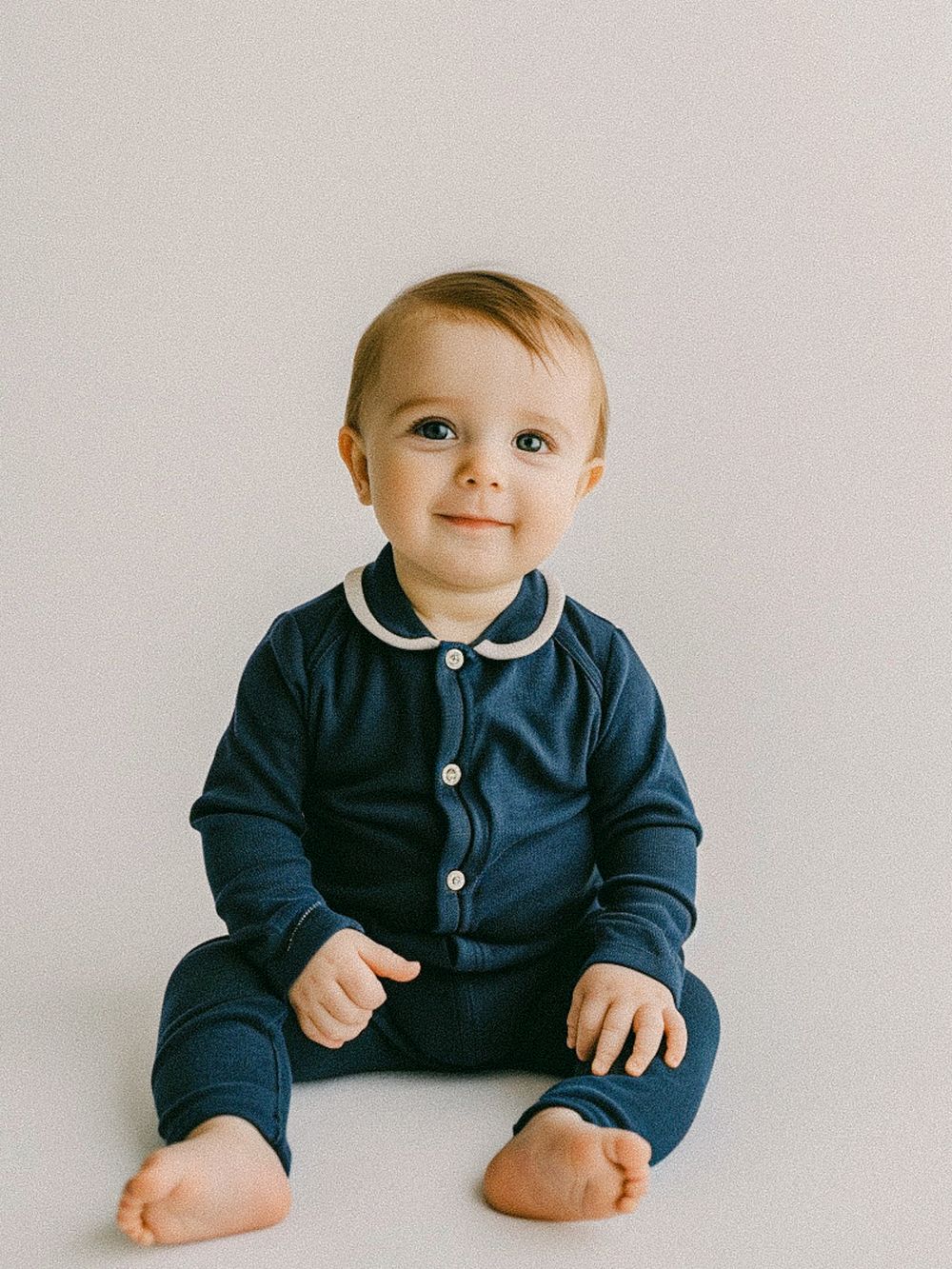 Baby wearing navy blue outfit with white collar sitting on floor.