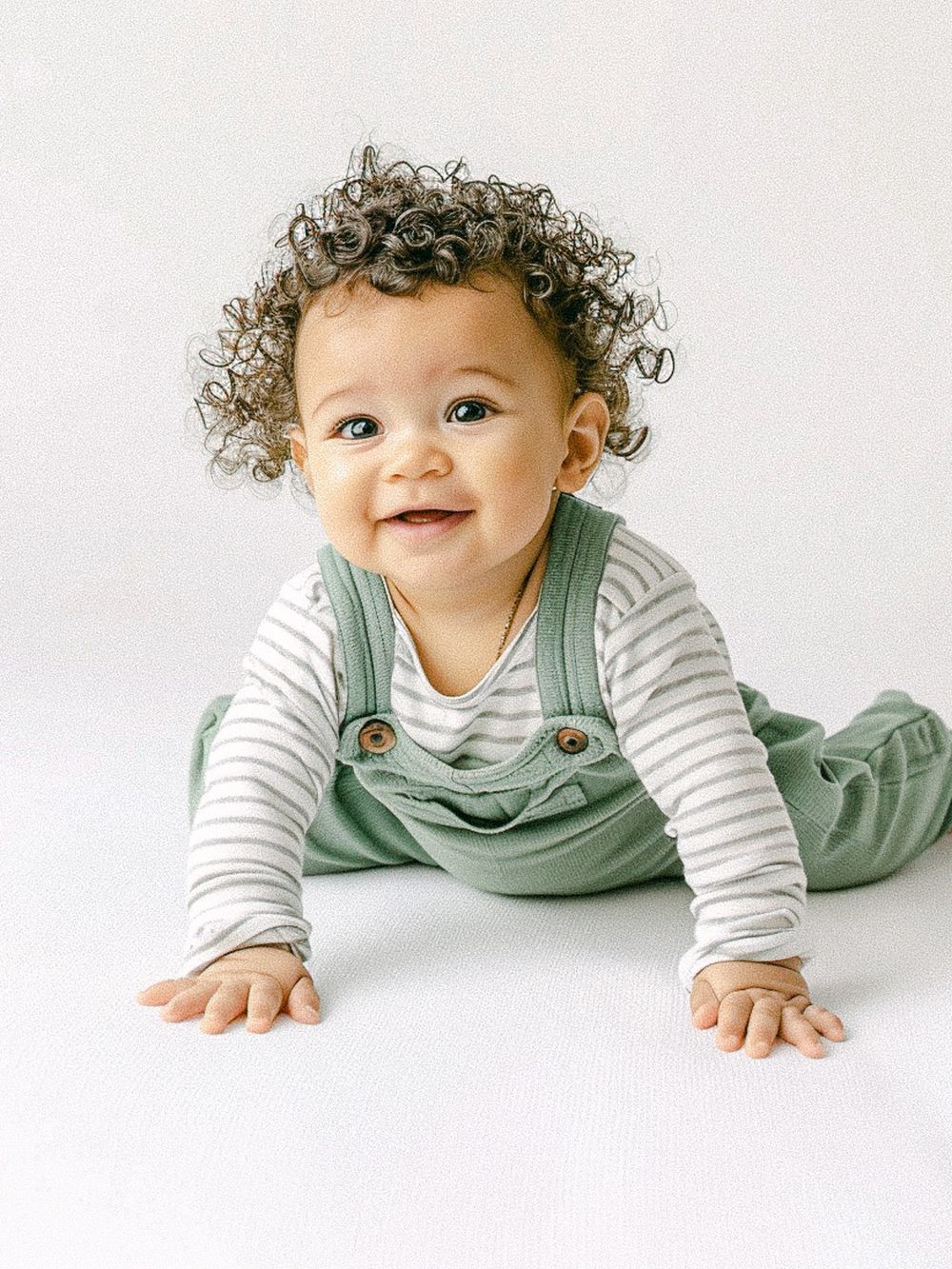 Un bébé souriant, portant un t-shirt rayé et une salopette vert menthe, aux cheveux bouclés, rampant sur un fond blanc.