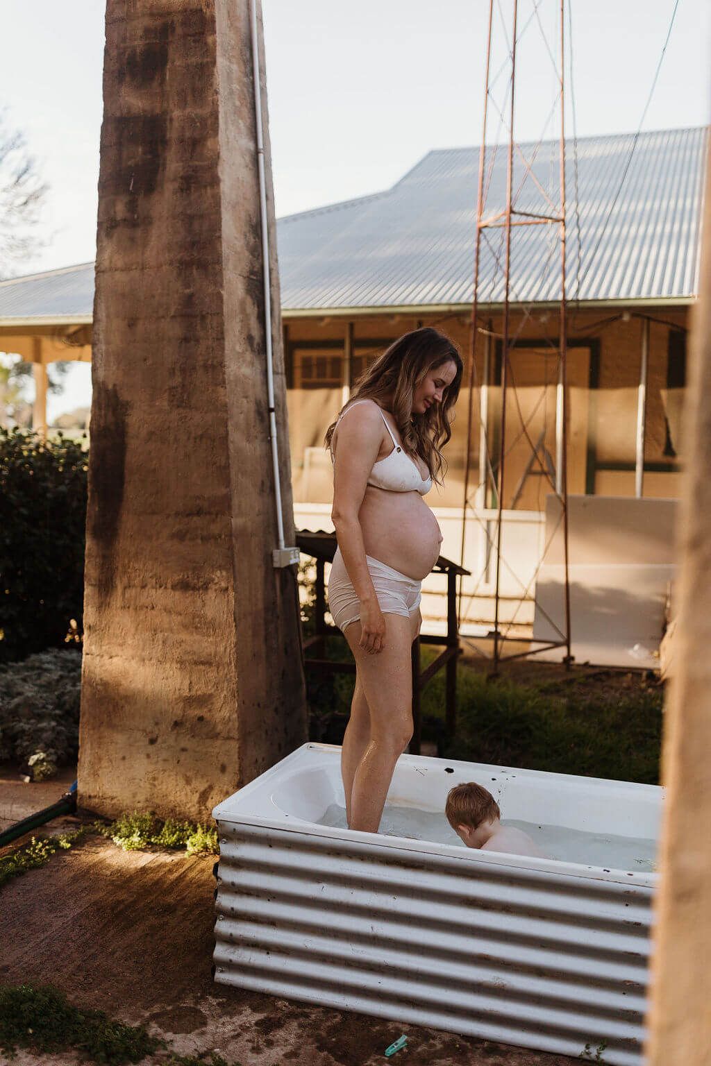 Pregnant mum standing in outdoor bath looking down at toddler son