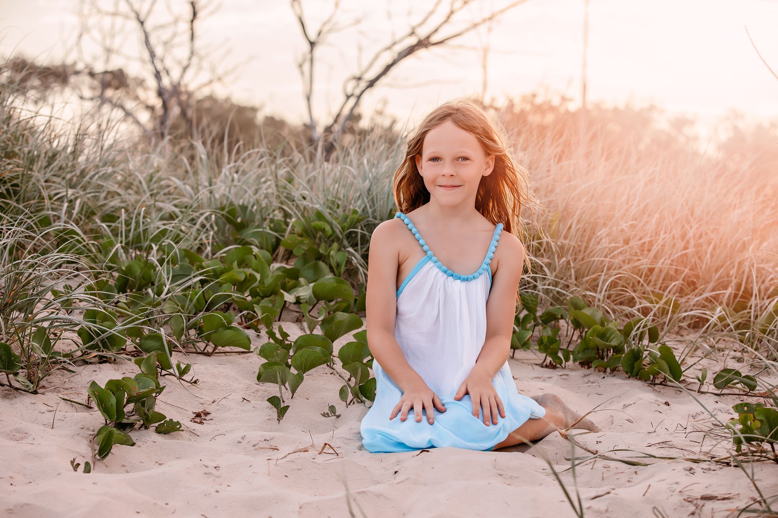 A young person in a light blue dress sits on sandy beach during golden hour.
