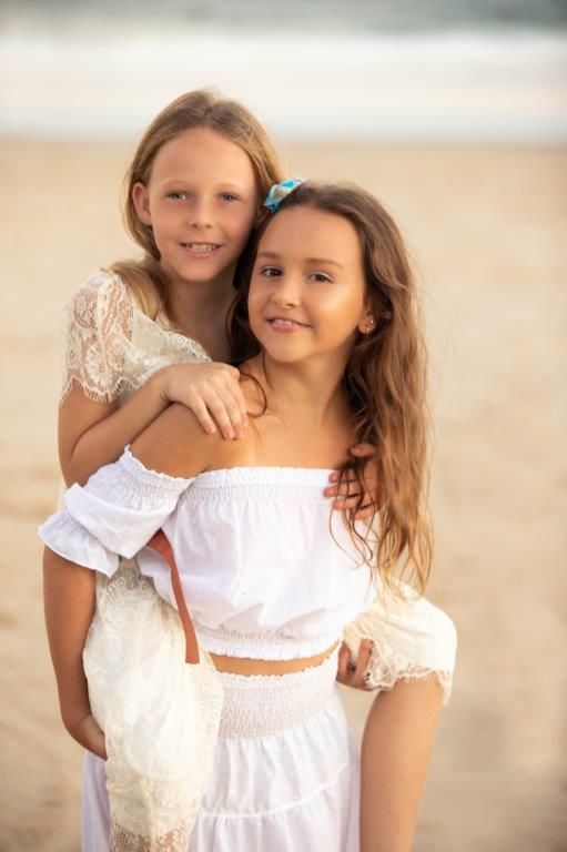 Two young girls in white dresses embracing on beach at golden hour.