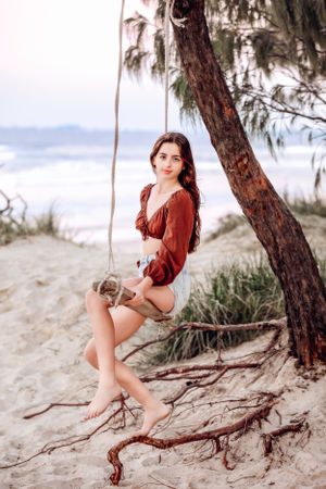 Person in red outfit posing on beach tree roots near ocean.