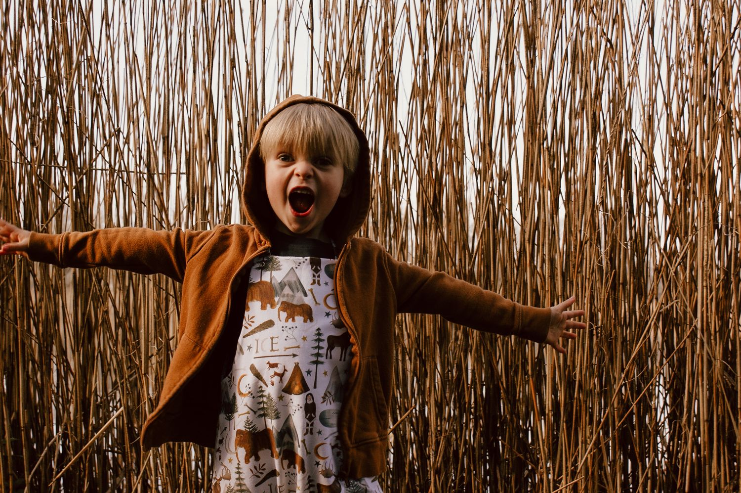 Child spreads arms wide while wearing  brown jacket and patterned dungarees against tall dried reeds background.